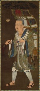 Tang dynasty Buddhist monk Xuanzang as a pilgrim. He carries a large backpack, which has a sun roof, and from that dangles a small incense burner. He holds objects in his hands, and wears sandals.