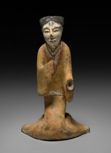 Painted clay figure of a kneeling woman. She appears to have a slight smile. One hand crosses over her chest, the other extends forward, the hand itself is missing.