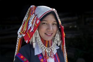 Photograph of a woman wearing a Hani costume. The headdress is a white hood with multicoloured sides, and a cord with tassels loosely draped under her chin.